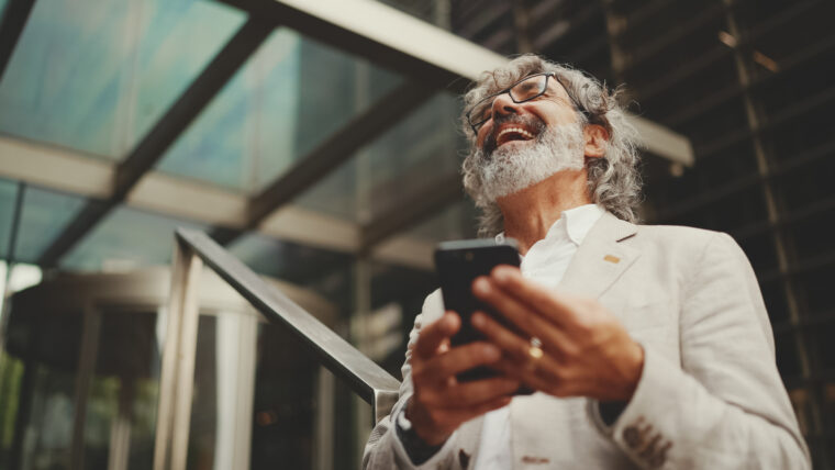 Mature businessman with beard in glasses stands on the street at the business center background. Middle aged manager texting message scrolling tapping to smartphone. Joy from good deal