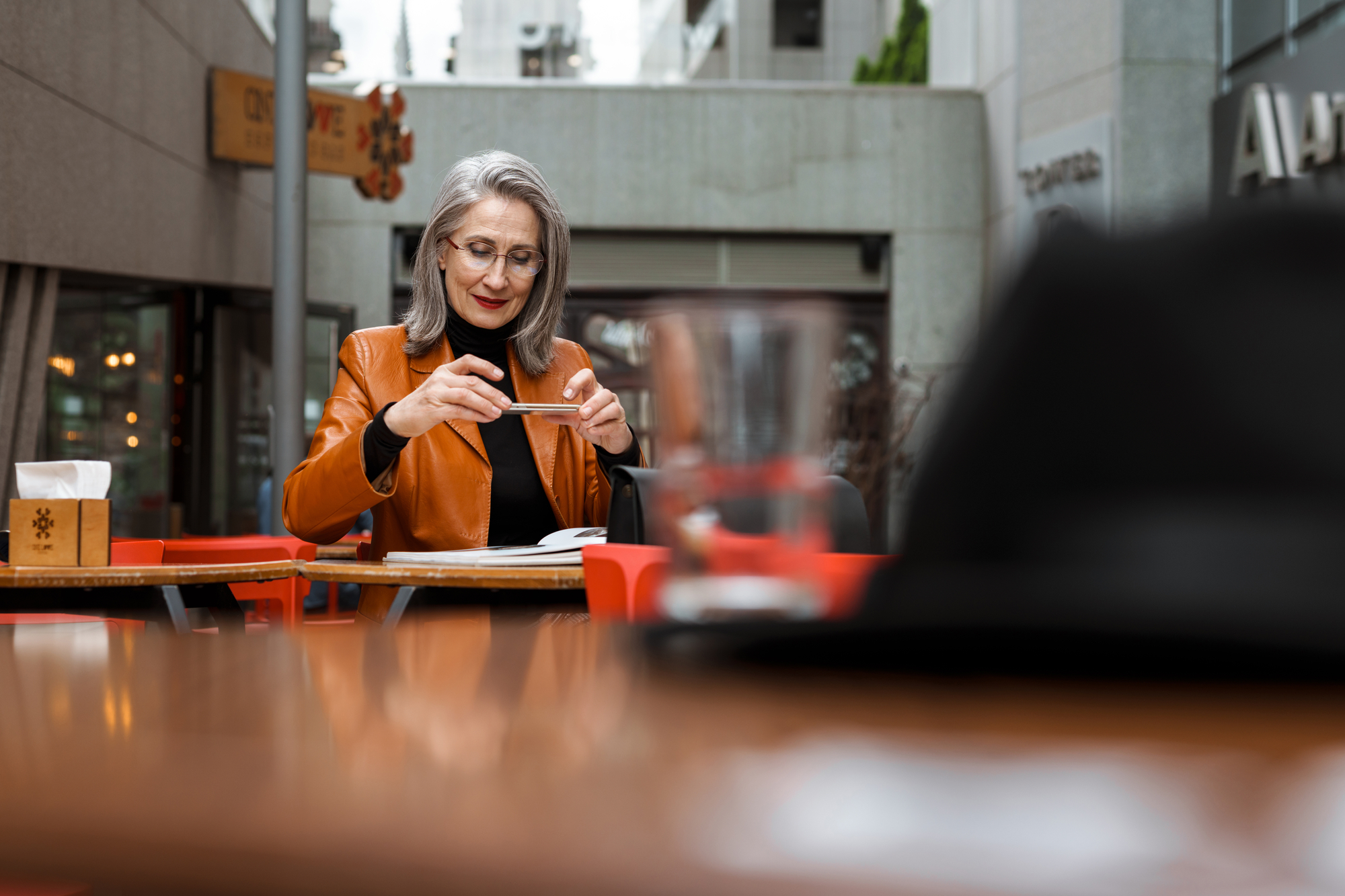 Grey white senior woman taking photo on cellphone while reading book in cafe outdoors