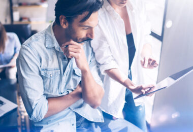 Coworking team meeting.Group of two businessmans working with new crafting startup project in sunny office.Touch pad in woman hands. Panoramic windows on blurred background.Closeup