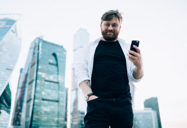 Half length portrait of happy male standing at urban setting with metropolis on background holding cellphone gadget and looking at camera, concept of millennial on city street