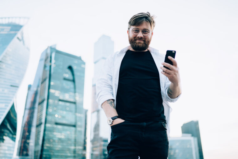 Half length portrait of happy male standing at urban setting with metropolis on background holding cellphone gadget and looking at camera, concept of millennial on city street